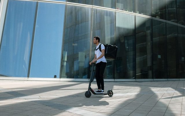 Commuter with a backpack riding a kick scooter past a glass building