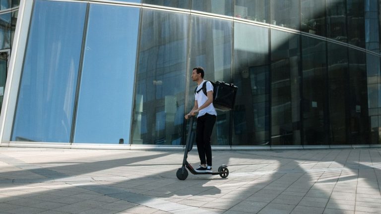 Commuter with a backpack riding a kick scooter past a glass building