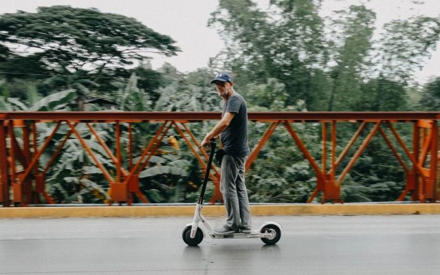 Man riding a shared electric scooter across a leafy bridge