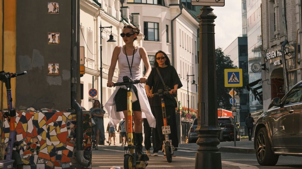 Two women riding electric scooters on a city sidewalk
