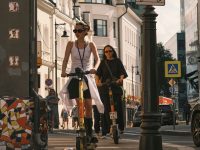 Two women riding electric scooters on a city sidewalk