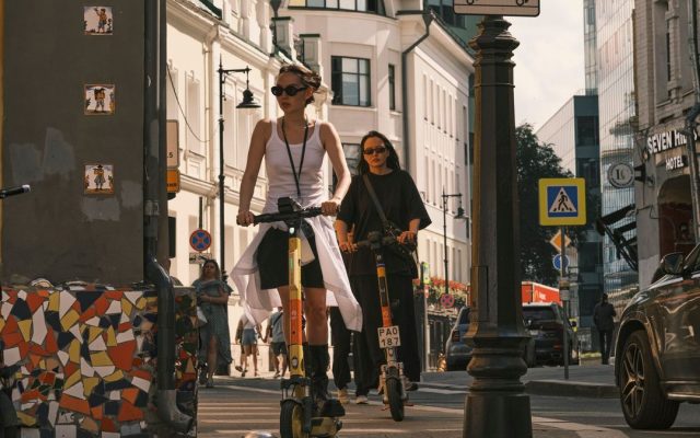 Two women riding electric scooters on a city sidewalk