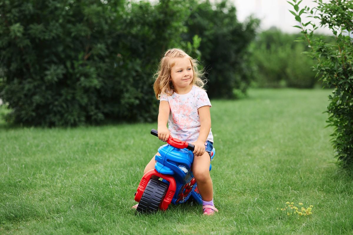 A cute girl in casual clothes rides a colorful toy bike on a green lawn