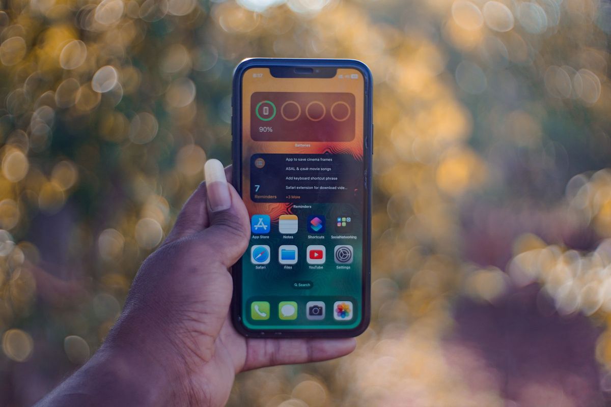 A hand holding a smartphone showing home screen apps, outdoors with bokeh background