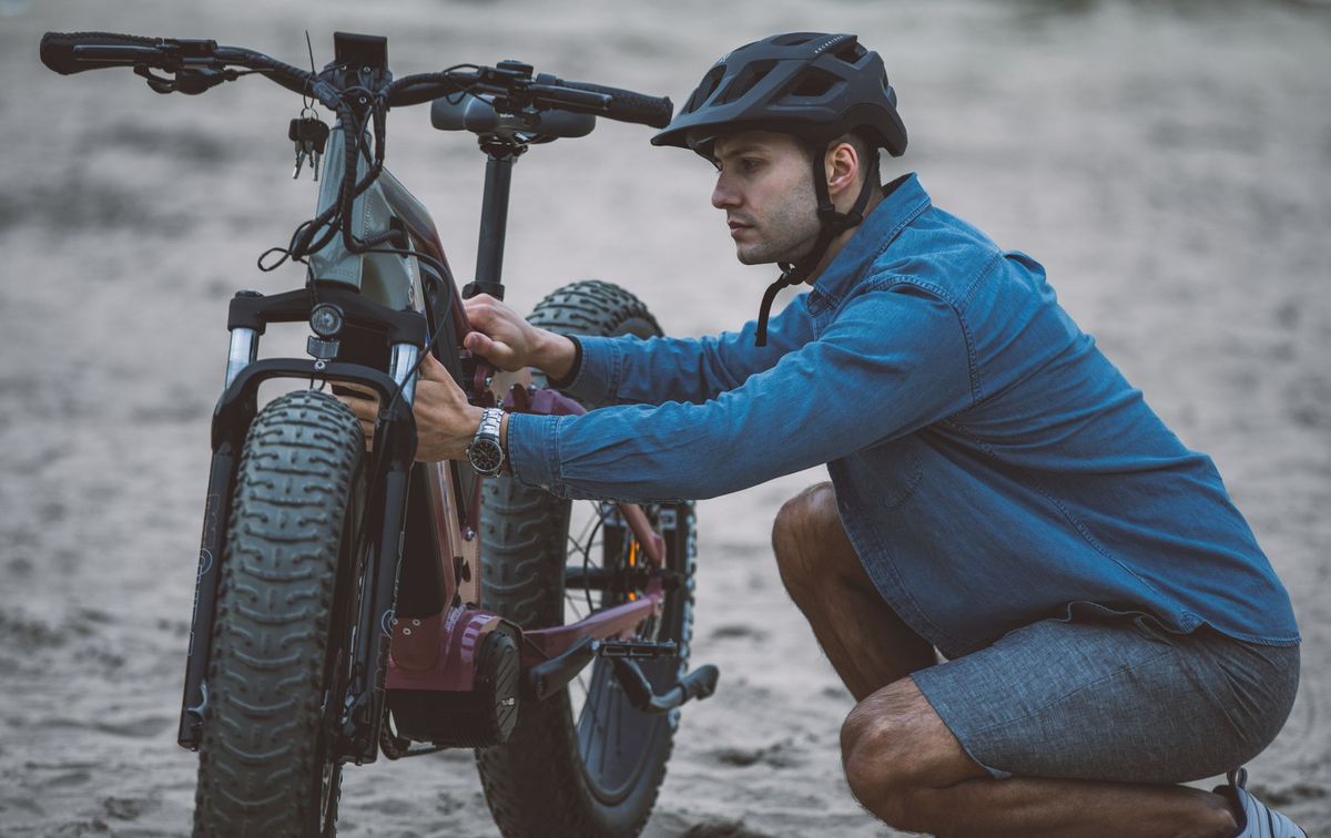 A man in a blue shirt and helmet fixing an electric bicycle on sandy terrain