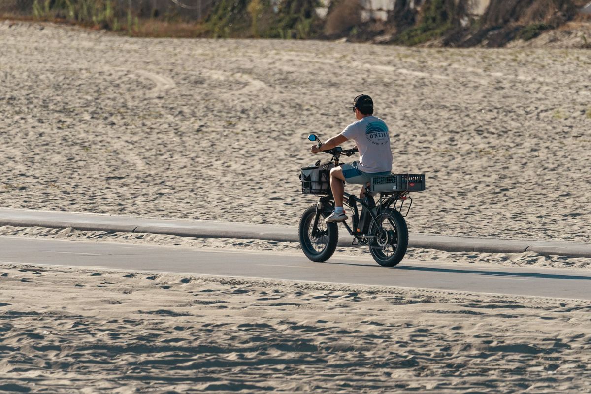 A man rides an electric bike along a sandy beach path under a clear sky, embracing outdoor adventure