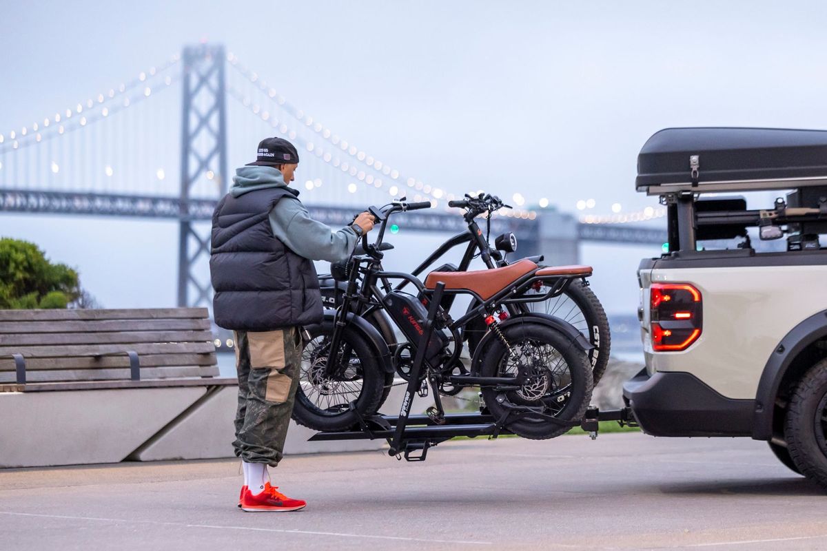 A man secures electric bikes onto a vehicle beside a lit suspension bridge at dusk