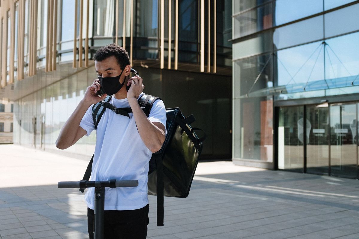 A man wearing a face mask and backpack rides an electric scooter, providing food delivery service