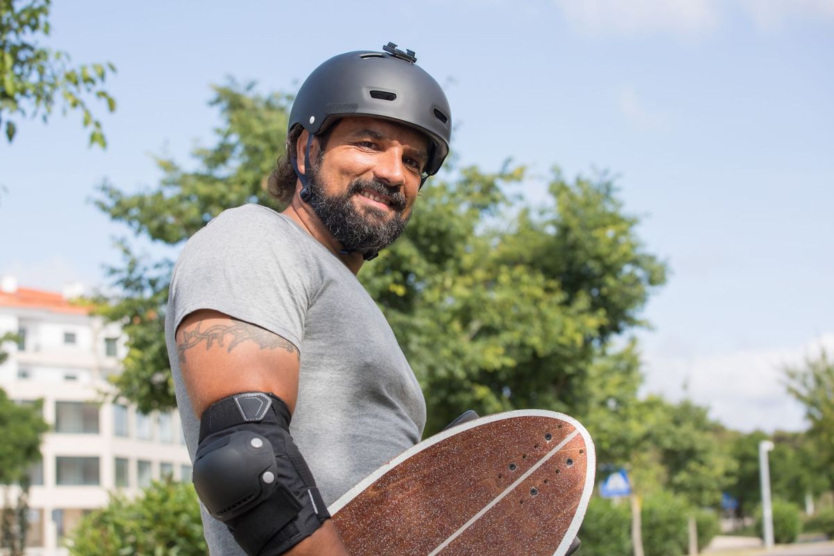 A man wearing a helmet and elbow pads holding a skateboard, smiling in an outdoor setting