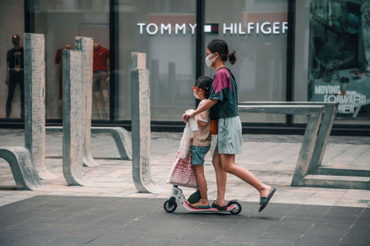 A woman and child ride an electric scooter in an urban environment, wearing masks