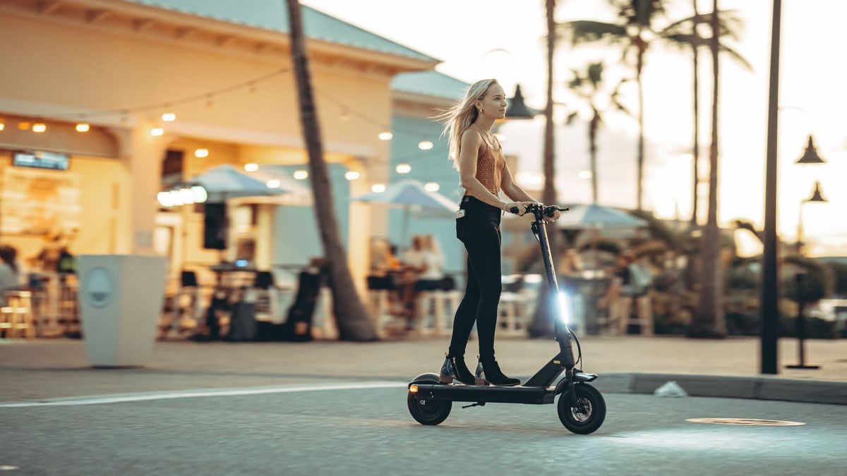 A woman enjoys a sunset ride on an electric scooter in a vibrant city setting with palm trees