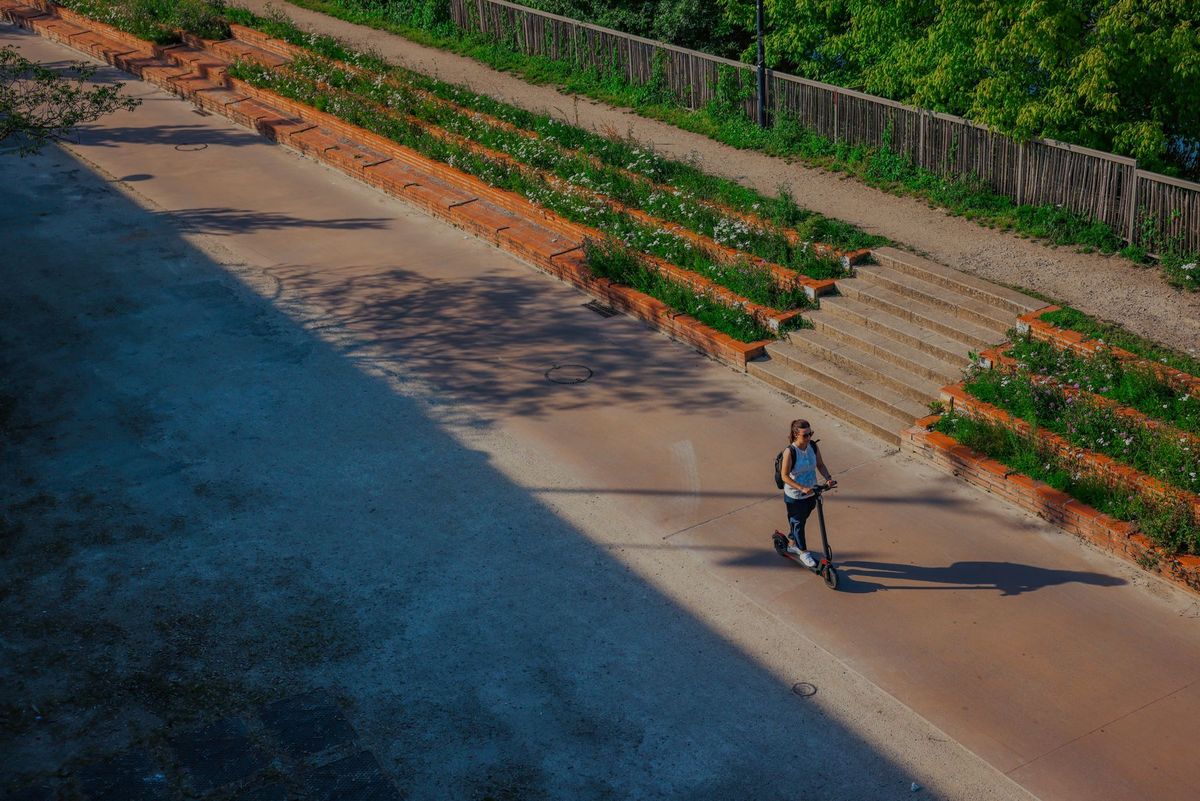 A woman rides an electric scooter on a sunny day in Bordeaux, France, near a park with greenery