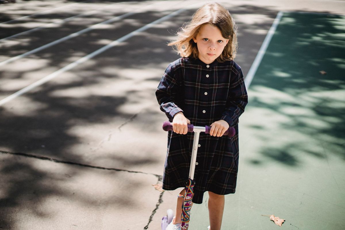 A young girl in a checkered dress rides a scooter on a sunny day at a playground