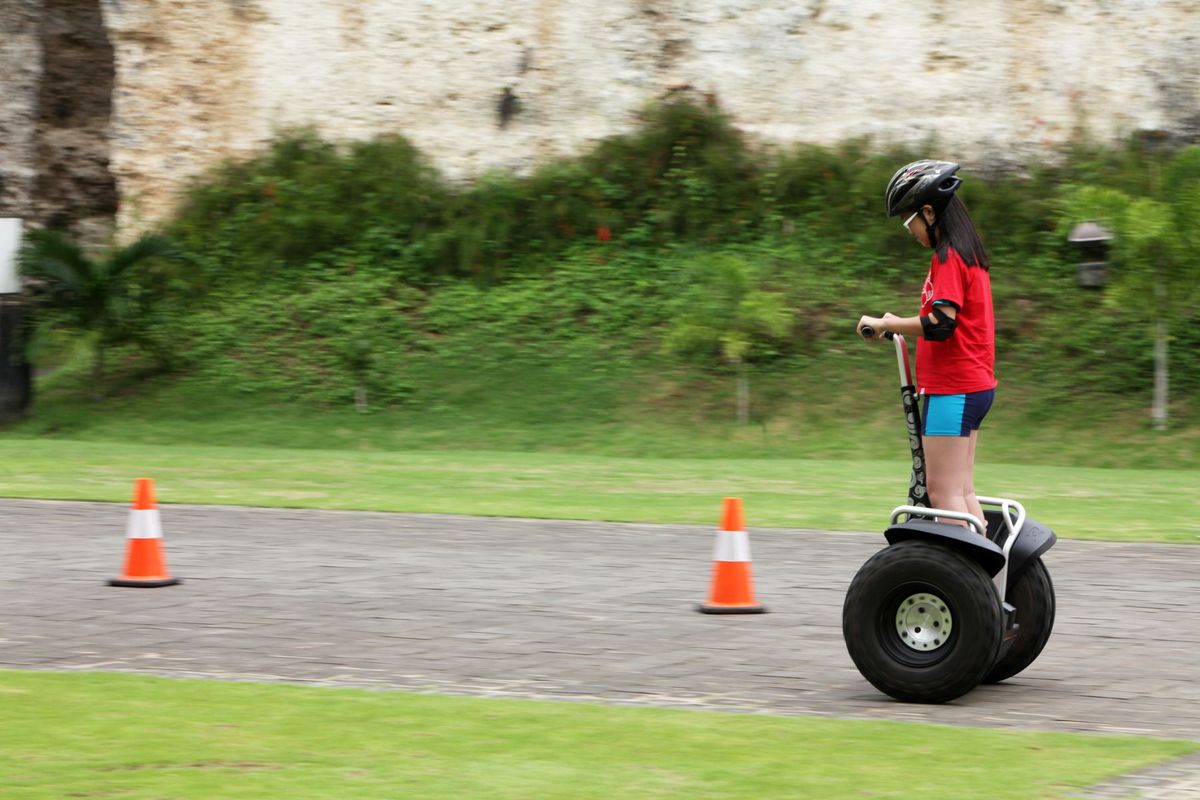 A young girl in safety gear riding a Segway on a paved path outdoors, surrounded by nature
