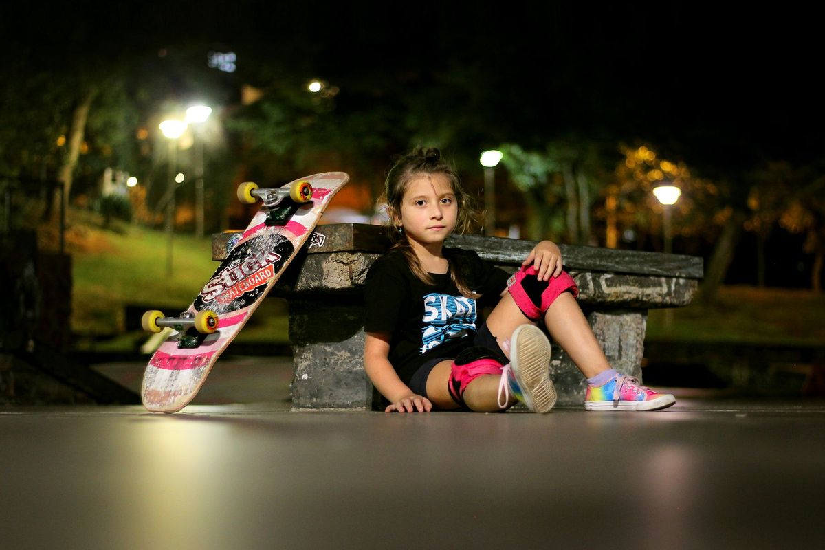 A young girl with a skateboard, sitting at night in Porto Alegre, wearing knee pads