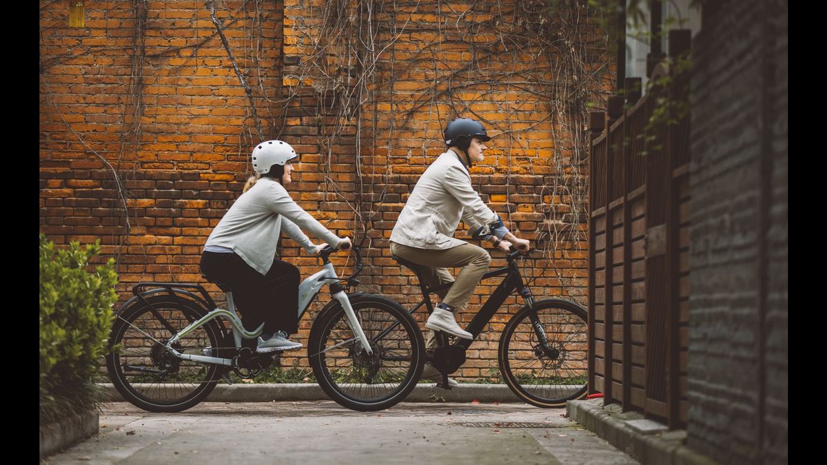 Couple cycling on E-bikes through a city lane with a classic brick wall backdrop