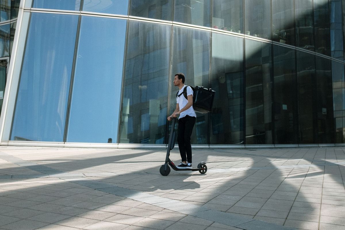 Man on electric scooter with backpack in front of modern glass building, representing urban delivery service