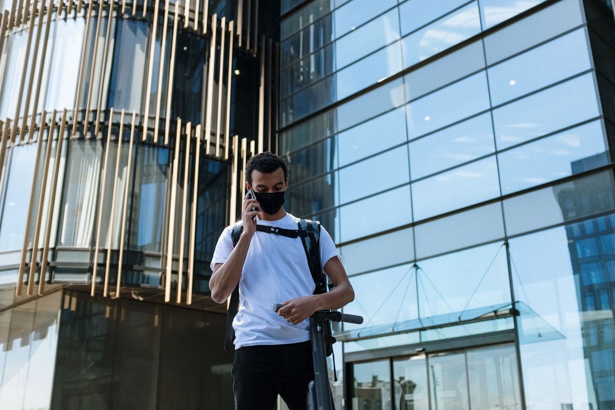 Man with black mask and backpack stands with an electric scooter in a modern city setting