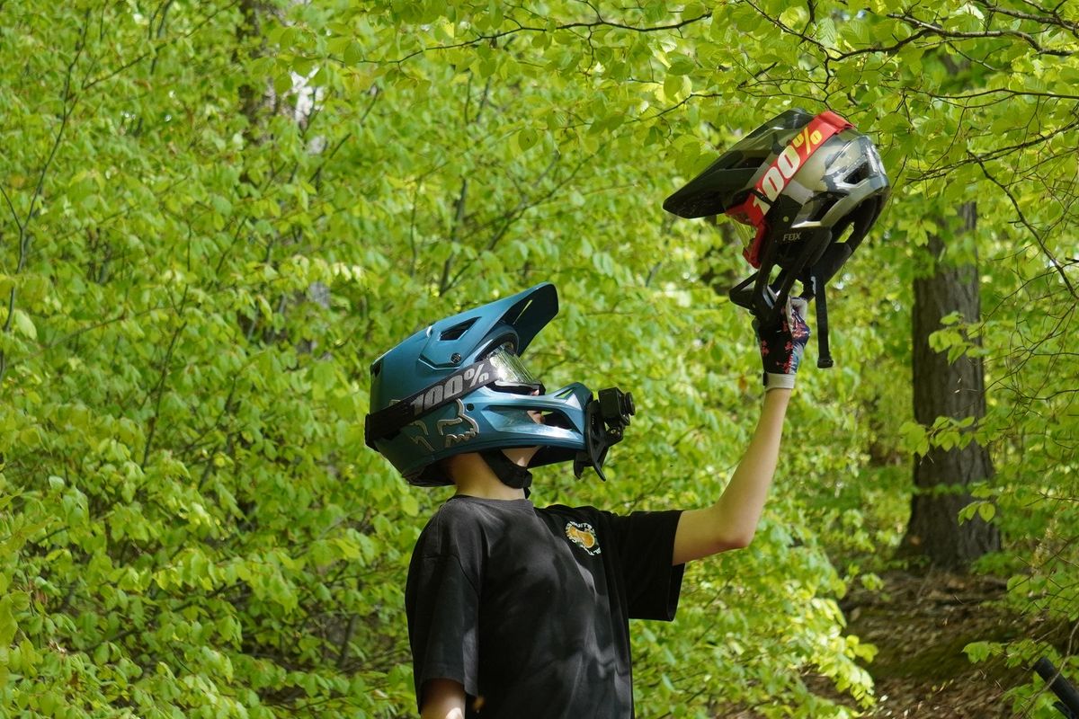 Mountain biker in forest holding one helmet while wearing another, surrounded by lush green foliage