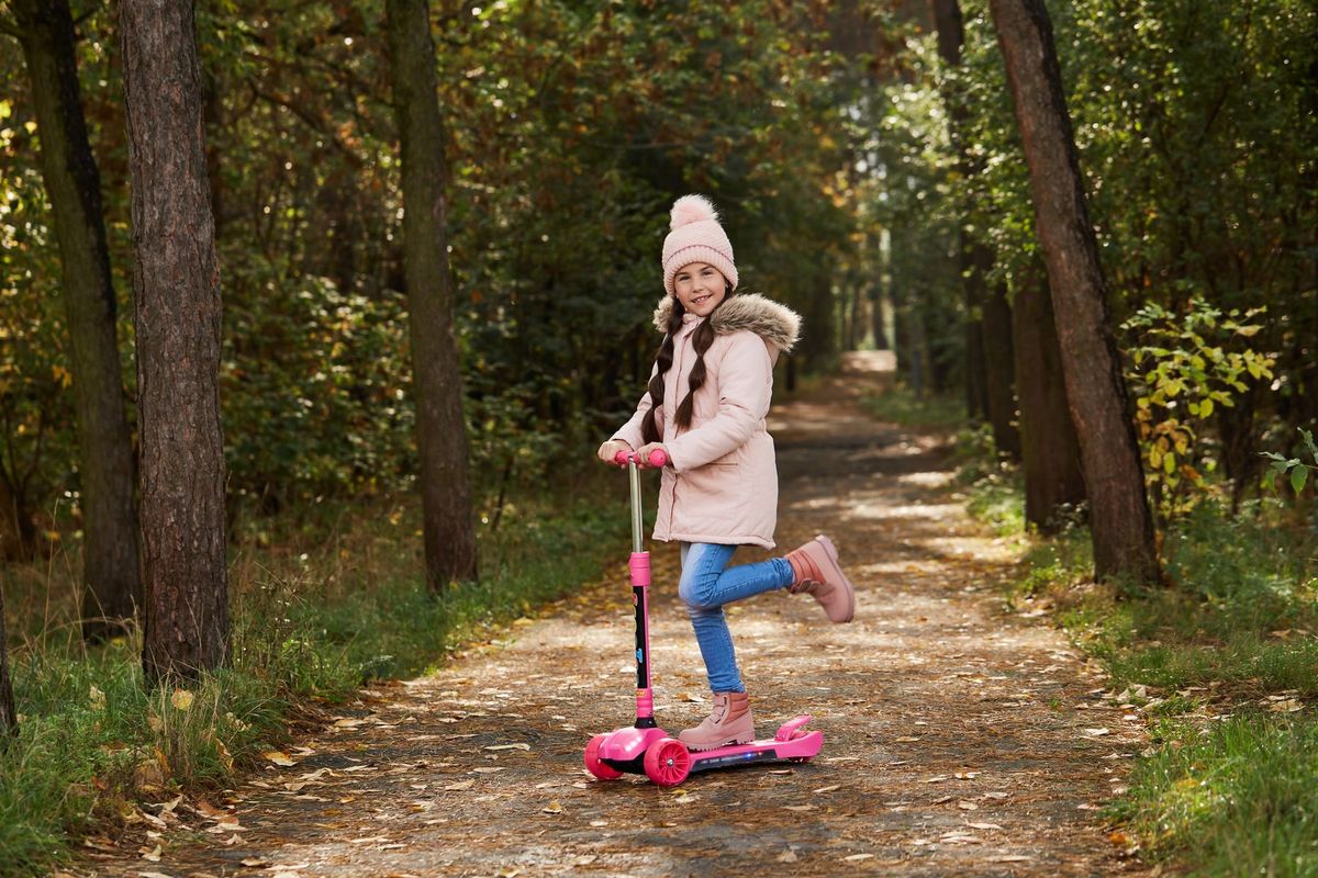 Smiling girl with beanie riding a scooter on a tree-lined path in autumn