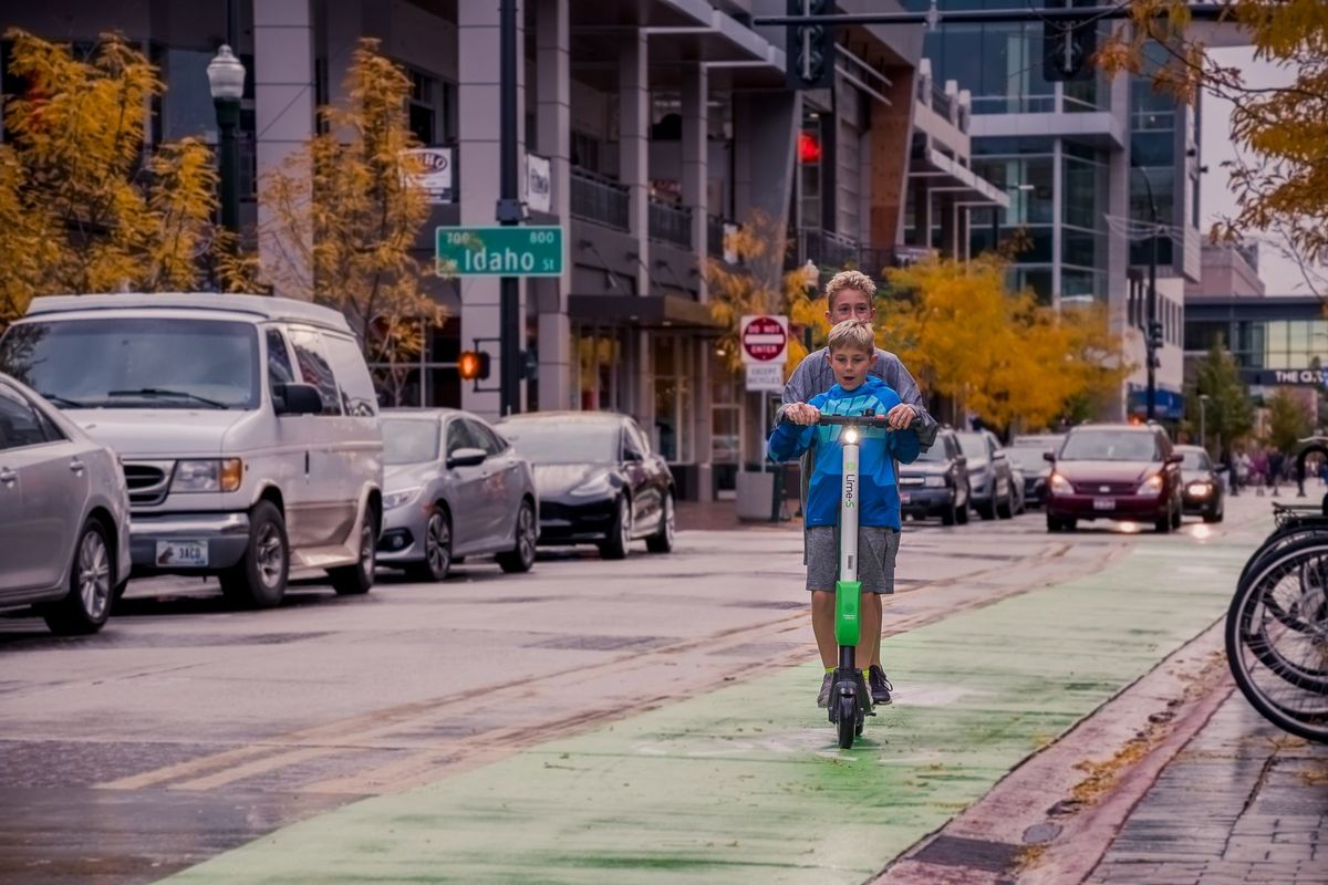 Two boys riding an electric scooter on a city street with autumn trees and traffic