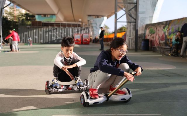 Two children riding hoverboard go-kart attachments in a skate park