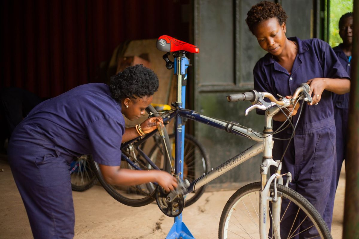 Two female mechanics in blue overalls working on a bicycle inside a workshop