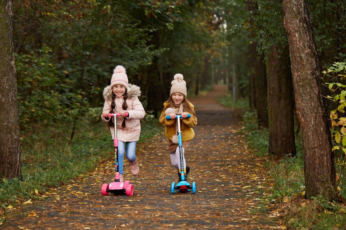 Two girls wearing beanies and winter jackets ride scooters on a leaf-strewn path in an autumn park