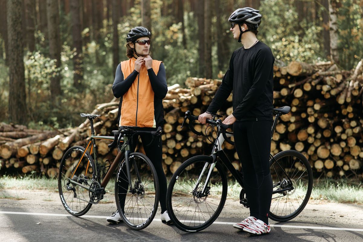 Two male cyclists pause on a forest road beside stacked logs, wearing helmets and athletic gear