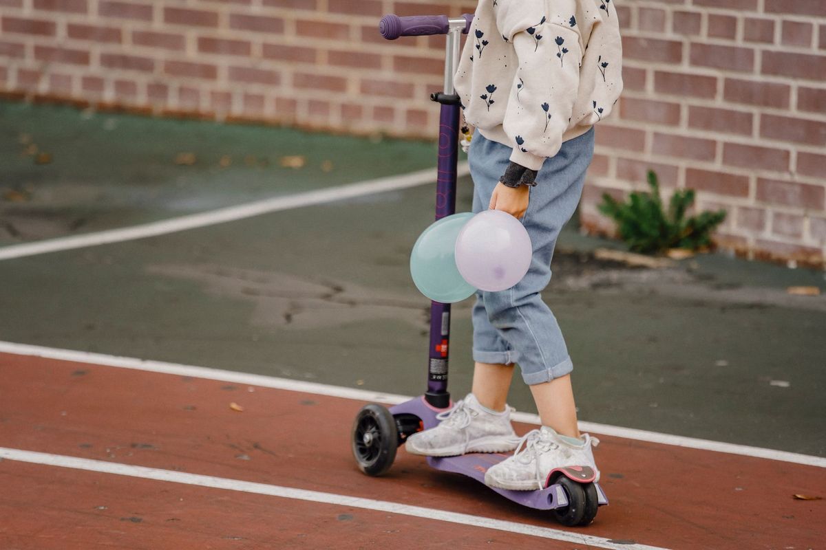 Young child riding a kick scooter holding balloons in an urban setting. Side view